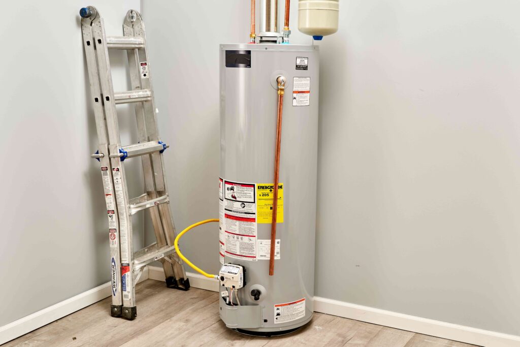 Tall gray water heater with copper pipes and warning labels in a utility room, next to a leaning metal ladder
